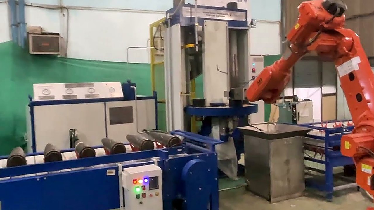A delivered bomb shell hydraulic pressure testing cell at work: shell bodies lying in cradles along a blue slat conveyor, the hydraulic test bed with its gauge panel behind, and the handling robot reaching in to load the station