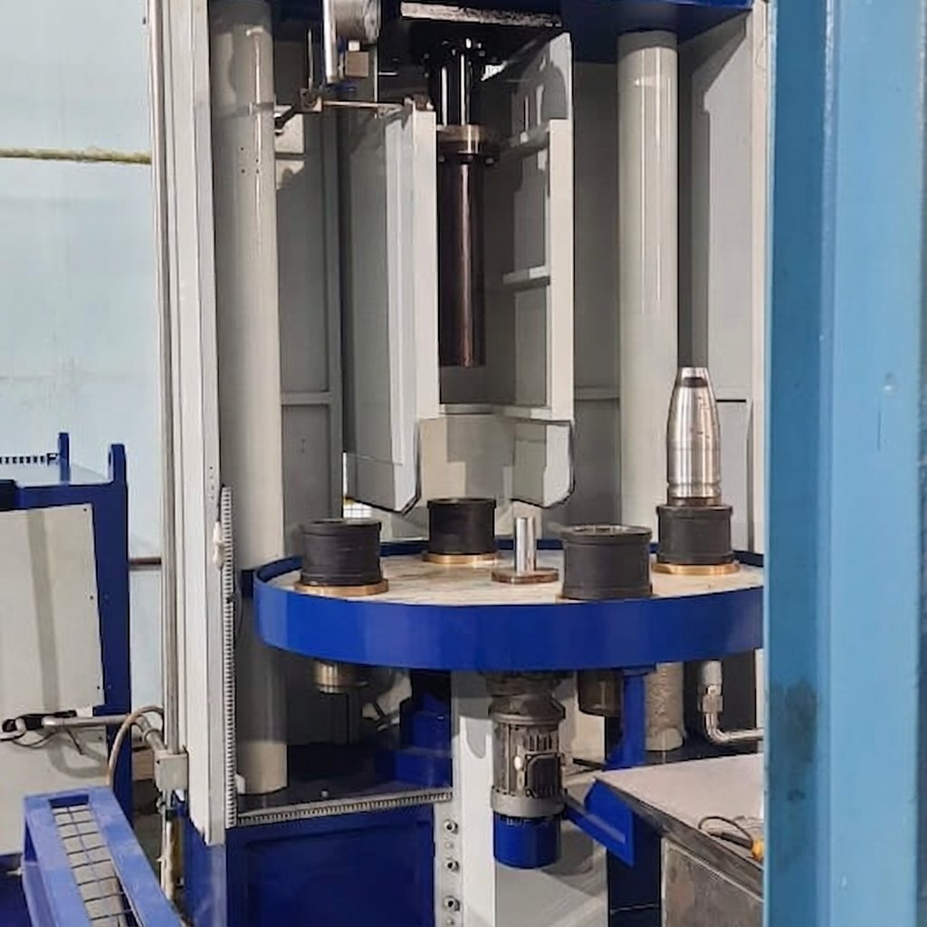 Photograph — delivered Neometrix rotary-carousel shell pressure-testing machine: blue frame, grey columns, rotary table with tooling rings and a single machined nose cone, at a customer works