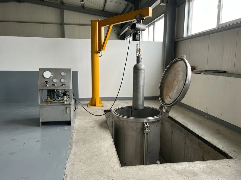 Illustrative image, not a delivered installation — the hydro test bay of a cylinder requalification facility: a vertical water-jacket test vessel set into the floor with its lid open, a plain steel air bottle suspended above it from a small hoist, a hydraulic power pack and gauge panel beside it, in a clean workshop bay