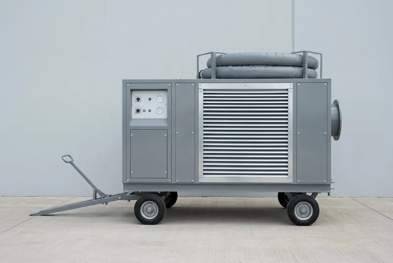 Illustrative of the class — a mobile air-conditioning trolley standing on empty clean concrete hardstanding: a boxy mid-grey painted sheet-metal enclosure on a welded steel chassis with four small solid rubber-tyred road wheels, a long steel tow drawbar angled down at the front, wide bare-metal louvred grilles across one side and the top for condenser air, a recessed blank control panel with plain unmarked round gauges, a large circular flanged air outlet at the opposite end and a coiled grey insulated flexible duct stowed in a cradle on top, no people, nothing running and no readable markings
