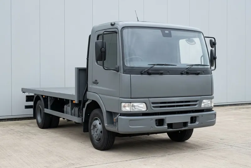 Illustrative of the class — the front three-quarter view of a plain matte mid-grey heavy fleet truck with a flat-fronted forward-control cab and an empty flat cargo bed, parked on empty concrete hardstanding: a small dark sensor pod mounted behind the top centre of the windscreen, a compact blank radar housing integrated low in the front bumper, and a slim black conduit running from the A-pillar toward the chassis, plain unmarked grille and an empty number-plate recess, no people, nothing running and no readable markings