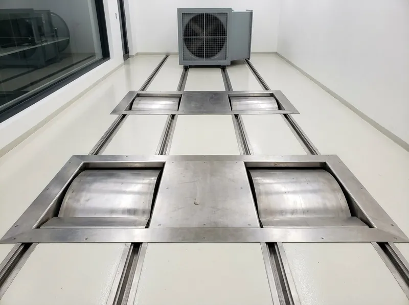 Illustrative image, not a delivered installation — a chassis dynamometer test cell: two pairs of large rollers set flush into stainless floor plates in a clean test bay, with centering rails between them, tie-down anchor points in the floor, a large vehicle cooling fan on rails at the front of the cell and a control room window along one wall