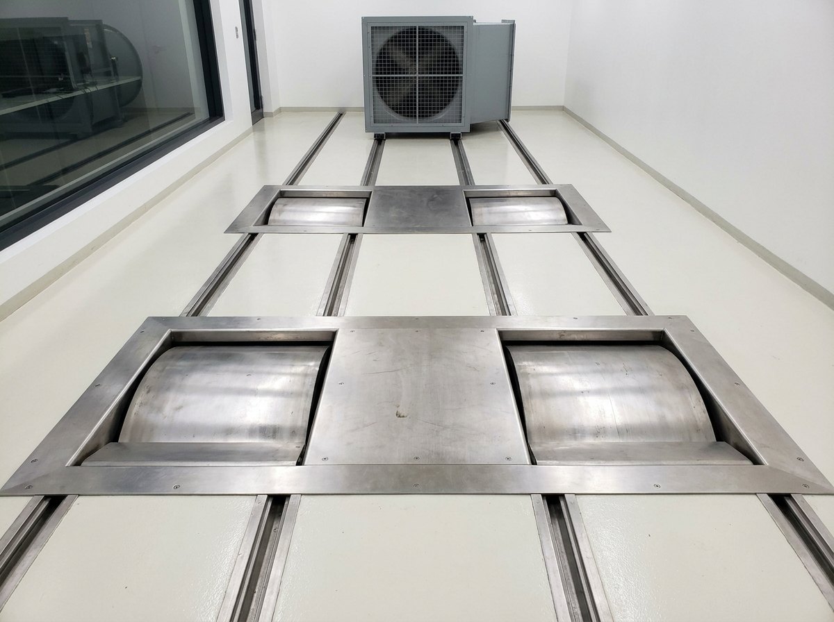 Illustrative image, not a delivered installation — a chassis dynamometer test cell: two pairs of large rollers set flush into stainless floor plates in a clean test bay, with centering rails between them, tie-down anchor points in the floor, a large vehicle cooling fan on rails at the front of the cell and a control room window along one wall