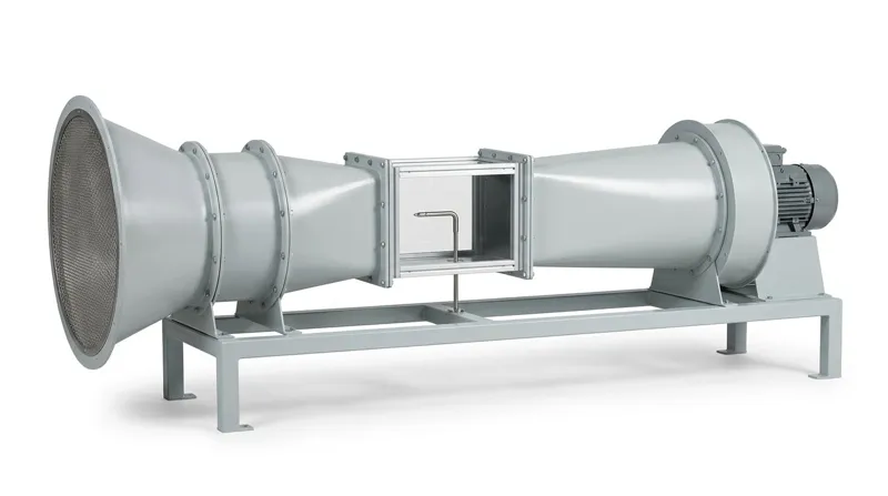 A calibration wind tunnel on a plain white studio background: a long horizontal tunnel of pale grey painted sheet steel on a low grey steel frame, with a wide bell-shaped inlet covered by a fine mesh, a flanged straight section, a tapering contraction, a clear test section in a bright aluminium frame with a slender pitot-static tube, a widening diffuser, and at the right end a round fan housing with a grey electric motor, and no people