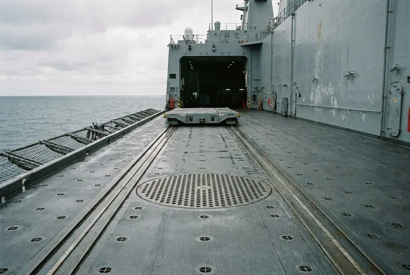 Illustrative image, not a delivered installation — the flight deck of a generic grey warship looking toward an open hangar: flush traversing rails running from a landing circle with a heavy perforated securing grid set into the deck, a low grey shuttle trolley parked on the rails, rows of tie-down points, no aircraft, no markings and no people