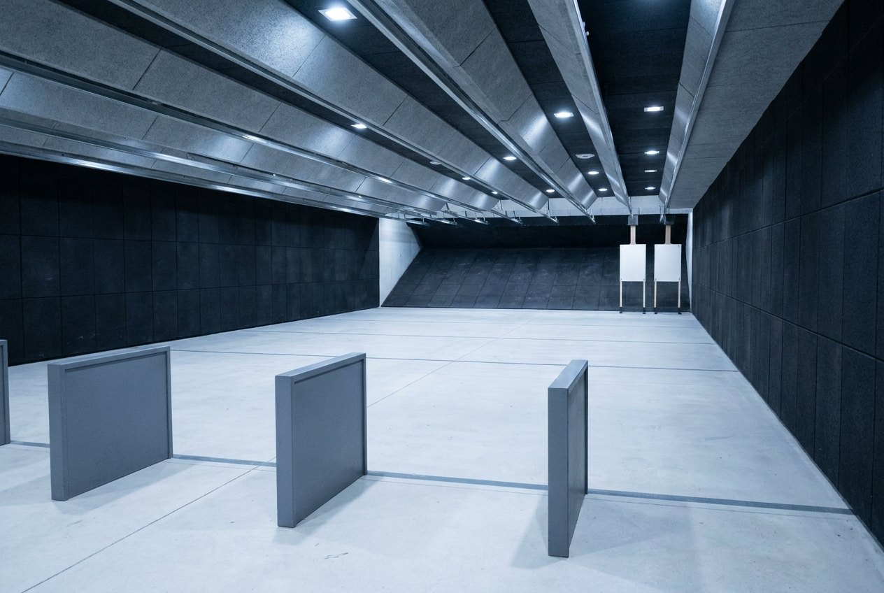 Illustrative image, not a delivered range — inside a modular indoor shooting range looking downrange: lit firing lanes with dividers, a baffled ceiling, dark rubber-lined walls, target frames at the far end before a rubber trap wall, clean floor with lane markings, no people, no firearms and no text