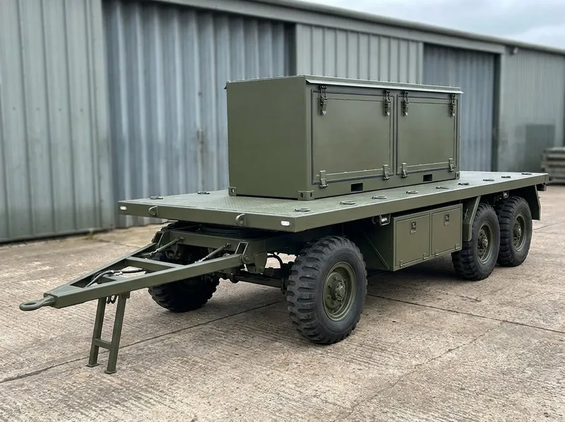 Munition handling trolley — olive drab three-axle flatbed trolley with steerable front axle, A-frame towbar and a latched rocket transport module on the deck