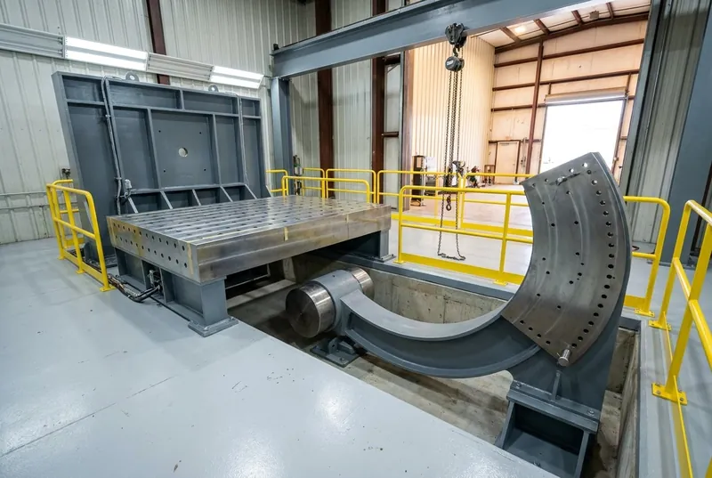 Illustrative of the class — a medium-weight-class naval shock qualification machine in a clean tall test hall: a heavy fabricated steel frame in fresh machine-grey paint standing over a shallow concrete pit, a very thick anvil table with a bright machined top drilled with a neat grid of tapped holes, a massive curved hammer arm with a cylindrical head resting at the bottom of its arc beneath the table, a tall curved steel quadrant plate carrying a row of plain drilled index holes and a locking pin, a chain hoist hanging from the frame beam, fresh yellow safety railing around the pit edge, clean light floor, no people and no readable markings