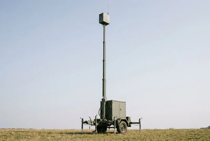 Illustrative image, not a delivered installation — a trailer-mounted telescopic mast extended to full height in an open field carrying a small plain generic payload box at the top, the trailer levelled on deployed outriggers with a control cabinet, no text, no markings and no people