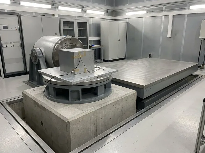 Illustrative image, not a delivered installation — an electrodynamic vibration shaker with a round armature table and a head expander, a generic boxy test specimen bolted on through a fixture with accelerometer cables, mounted on a seismic mass in a clean test laboratory, with a horizontal slip table beside it