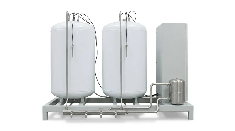 A water heater test facility on a plain white studio background: a low pale grey steel test frame holding two plain white enamelled cylinders side by side, a stainless steel pipe running down from each to a stainless manifold of four small valves along the front edge, thin black sensor cables with small stainless probe sleeves at the top of each cylinder, and at the right end a pale grey cabinet and a small stainless supply vessel joined to the manifold by two pipes, and no people